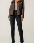A model wearing a brown leather jacket, black jeans, and black ankle boots, standing against a plain light beige studio background.