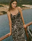 Woman in a black and white patterned dress standing on a wooden railing by a body of water with mountains in the background.