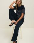 Woman sitting on a stool wearing a black and white striped shirt and dark pants against a plain background