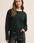 Woman wearing a dark green cardigan over a white shirt and black pants on a beige background