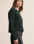 Woman wearing a green cardigan over a white shirt with black pants on a beige background