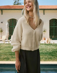 Woman wearing a cream sweater and black pants standing by a pool with a building in the background