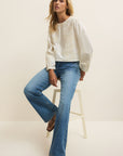 Woman wearing a white sweater and blue jeans sitting on a white stool against a plain background