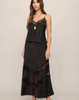 A woman modeling a black two-piece camisole and maxi skirt set with sheer lace panels, photographed against a plain light gray studio background.