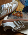 Pair of white sneakers with brown accents on a wooden surface with a painting in the background.
