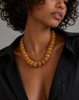 A close-up of a model wearing a chunky amber-brown beaded necklace with a black button-down shirt against a neutral gray background.