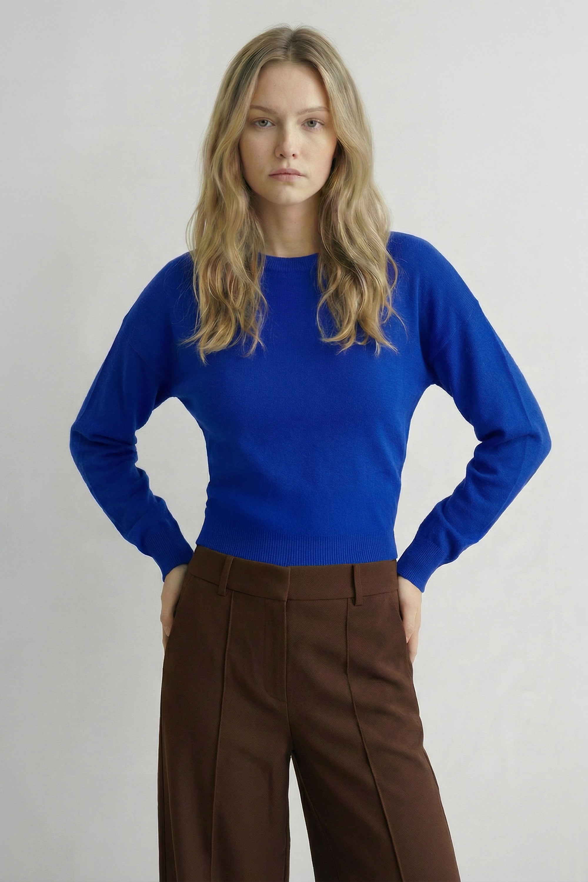 A woman modeling a bright royal blue long-sleeve knit top with brown trousers, standing against a light gray studio background.