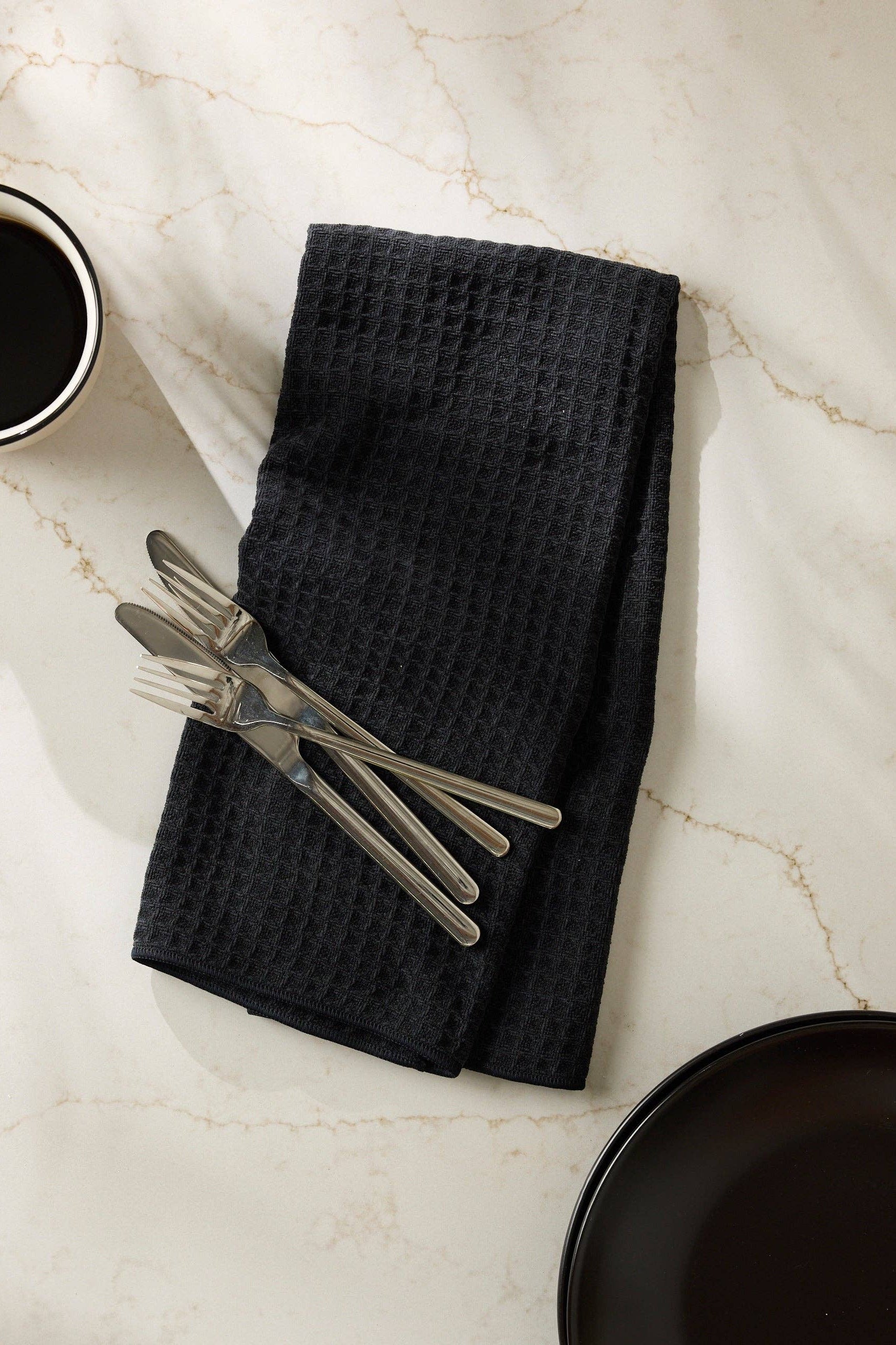 Black waffle-textured towel with silverware on a marble surface with a black plate and cup.