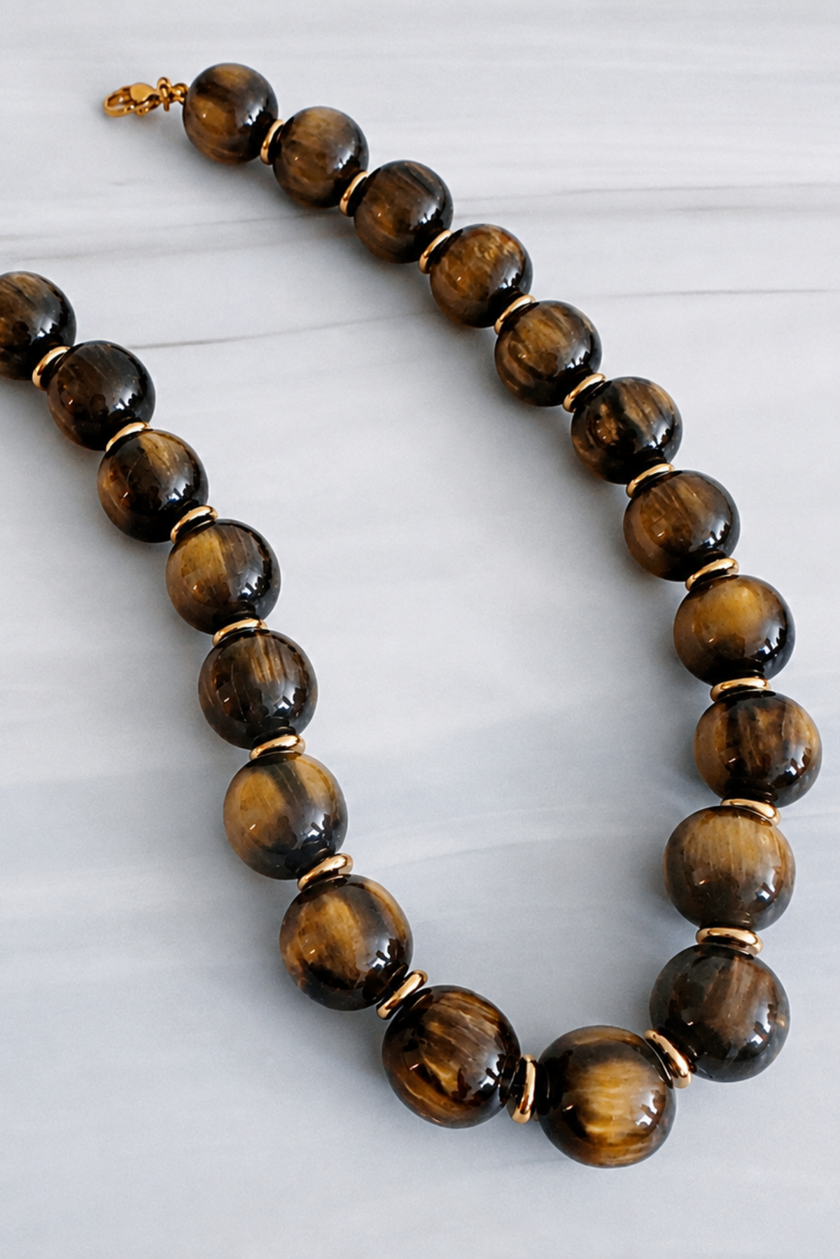 A beaded necklace made of polished tiger's eye beads with gold spacer rings and a gold clasp, displayed on a light gray marble surface.