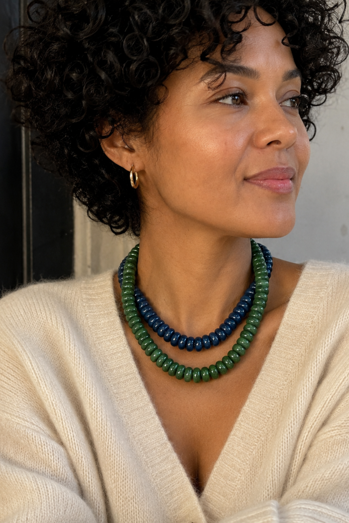 A woman with curly black hair wearing a cream knit sweater and a double-strand beaded necklace in green and dark blue, posed in a cafe setting.