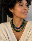A woman with curly black hair wearing a cream knit sweater and a double-strand beaded necklace in green and dark blue, posed in a cafe setting.