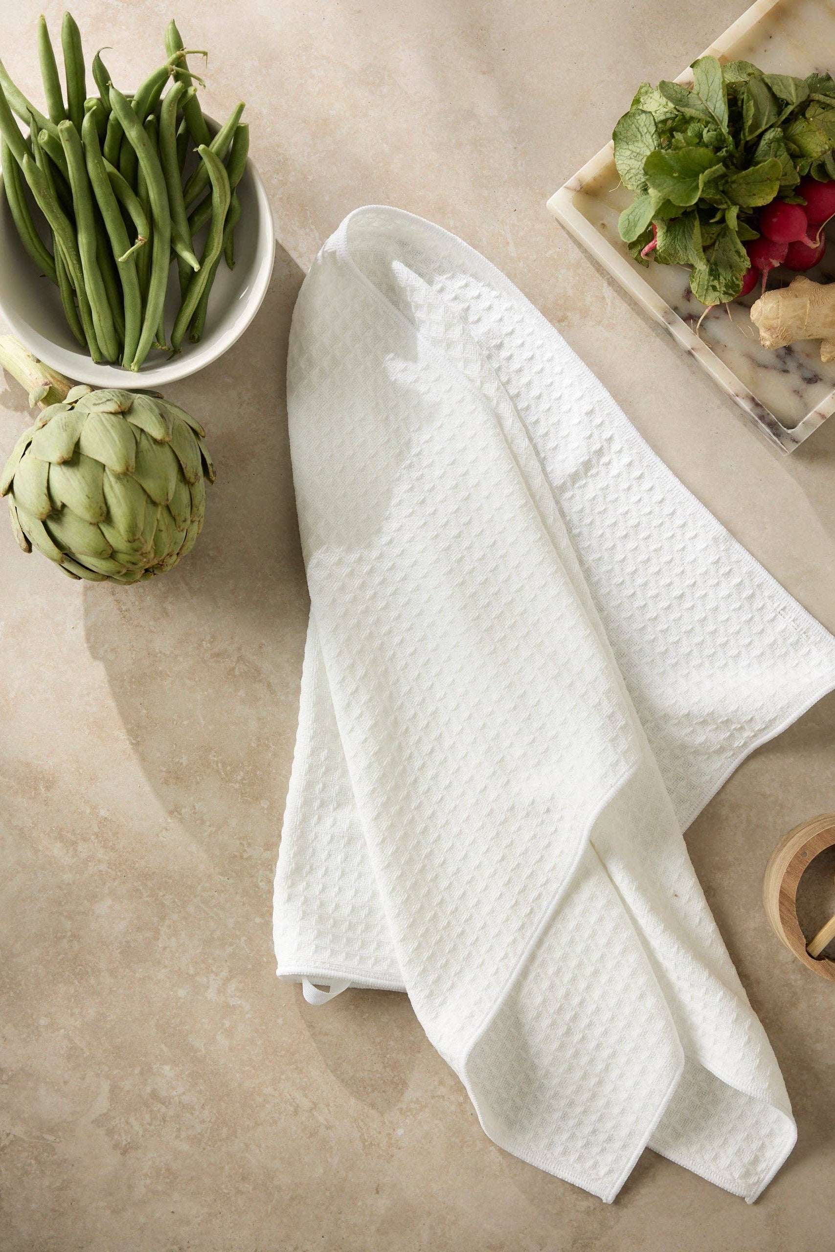 White kitchen towel on a beige surface with vegetables including green beans, artichoke, and radishes.