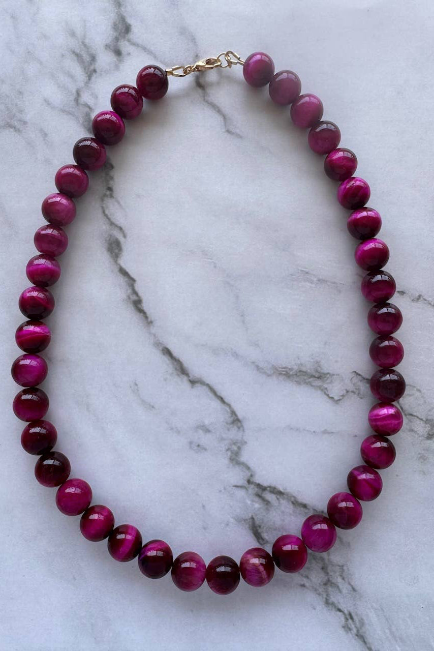 A beaded necklace made of glossy, round magenta-purple beads with a gold clasp, displayed on a white marble surface with gray veining.