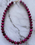 A beaded necklace made of glossy, round magenta-purple beads with a gold clasp, displayed on a white marble surface with gray veining.