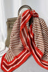 A red and beige geometric striped silk scarf with black dotted accents, draped over a taupe leather handbag on a gray surface with a white curtain backdrop.