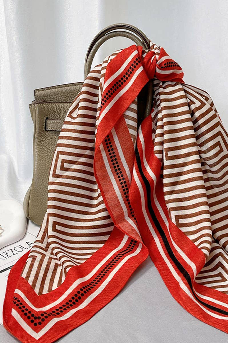 A red and beige geometric striped silk scarf with black dotted accents, draped over a taupe leather handbag on a gray surface with a white curtain backdrop.