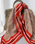 A red and beige geometric striped silk scarf with black dotted accents, draped over a taupe leather handbag on a gray surface with a white curtain backdrop.