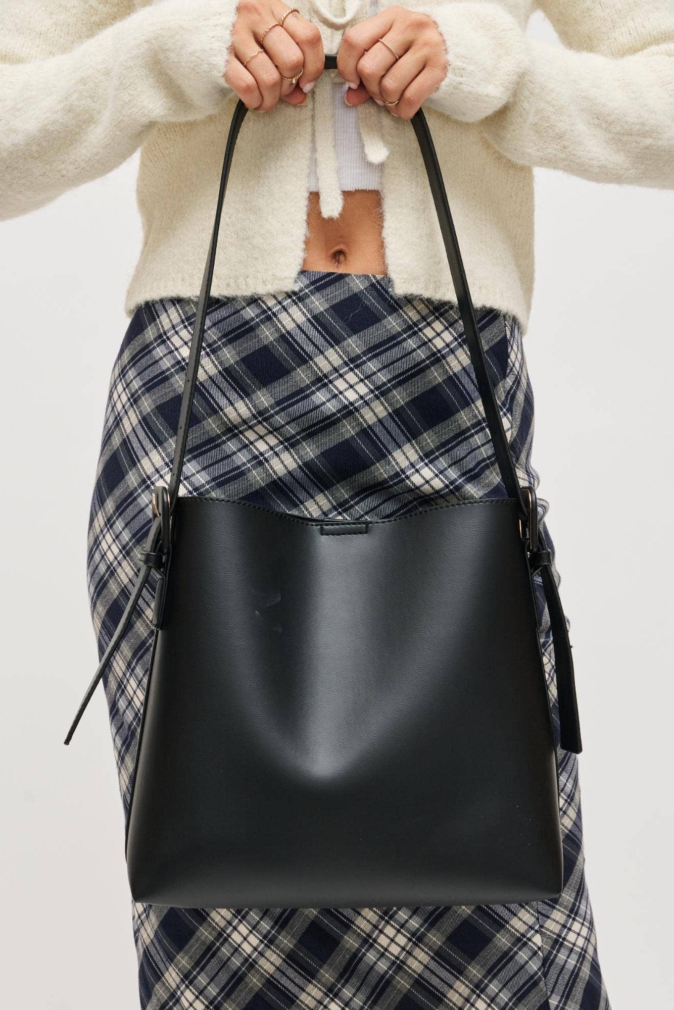 Person holding a black leather bag with plaid skirt and white sweater