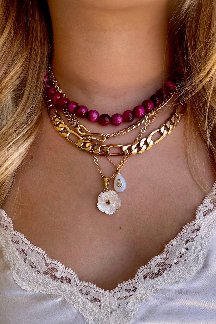 A close-up of a person's neck and chest wearing a layered necklace set. The set includes a magenta bead choker, a gold chain with a figure link and curb link, a gold chain with a white flower charm, and a gold chain with a teardrop shell charm.