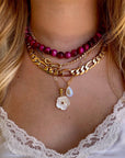 A close-up of a person's neck and chest wearing a layered necklace set. The set includes a magenta bead choker, a gold chain with a figure link and curb link, a gold chain with a white flower charm, and a gold chain with a teardrop shell charm.