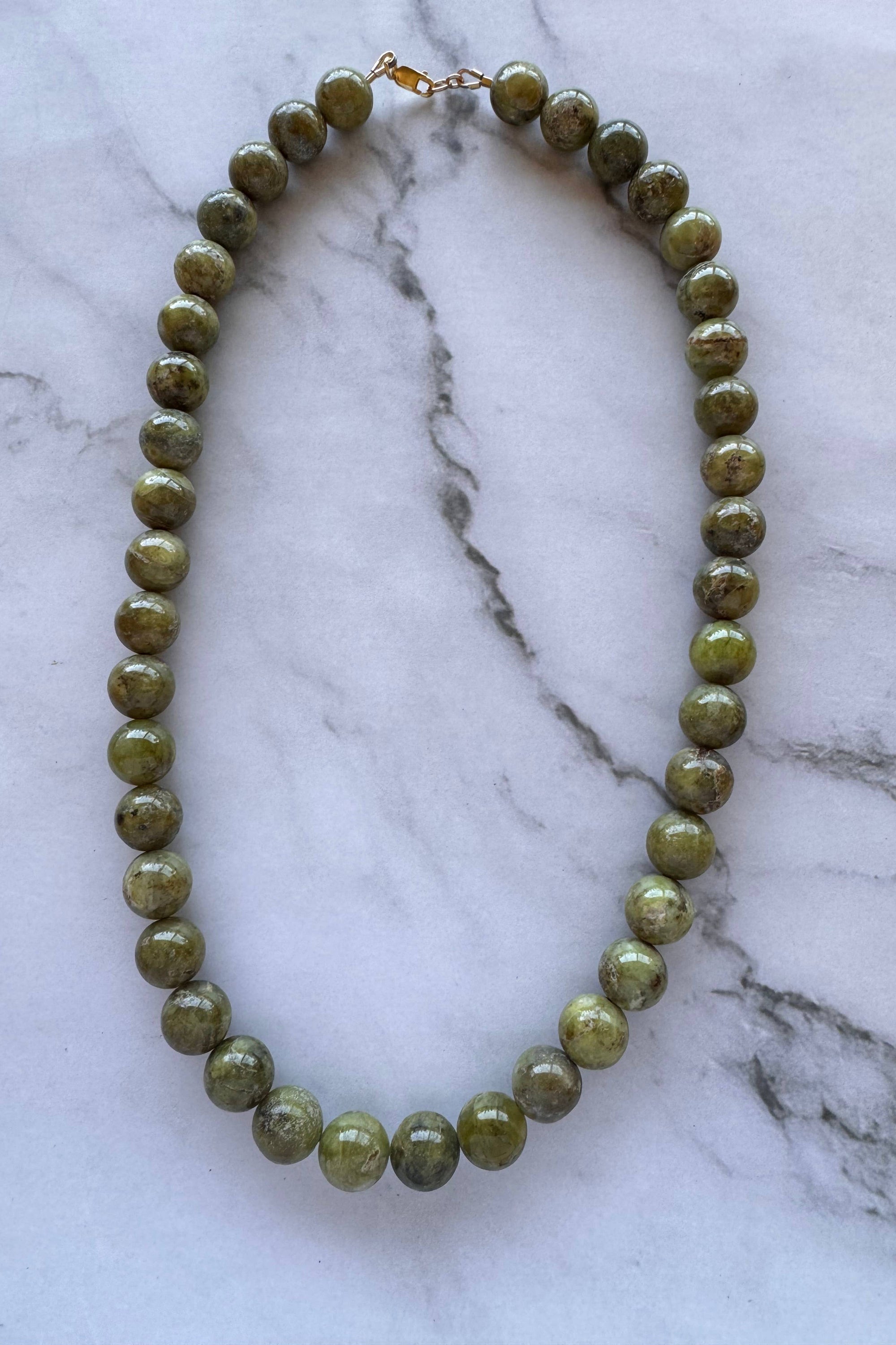 A beaded necklace made of polished olive green gemstone beads with a small gold clasp, displayed on a white and gray marble surface.