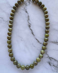 A beaded necklace made of polished olive green gemstone beads with a small gold clasp, displayed on a white and gray marble surface.