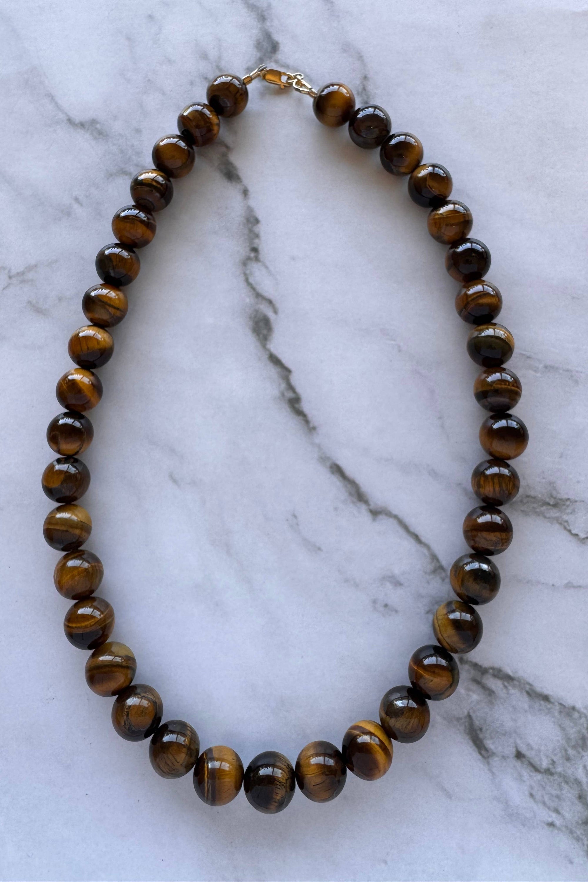 A beaded necklace made of polished brown tiger's eye beads with a small gold clasp, displayed on a white marble surface.