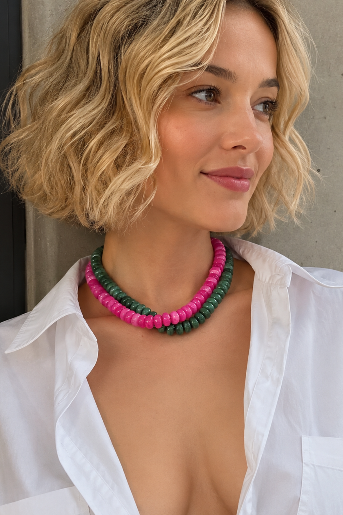 A woman with short blonde wavy hair wears a double-strand beaded necklace with pink and green beads, paired with an open white button-down shirt, posing against a concrete wall.