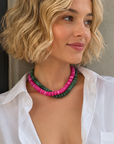 A woman with short blonde wavy hair wears a double-strand beaded necklace with pink and green beads, paired with an open white button-down shirt, posing against a concrete wall.