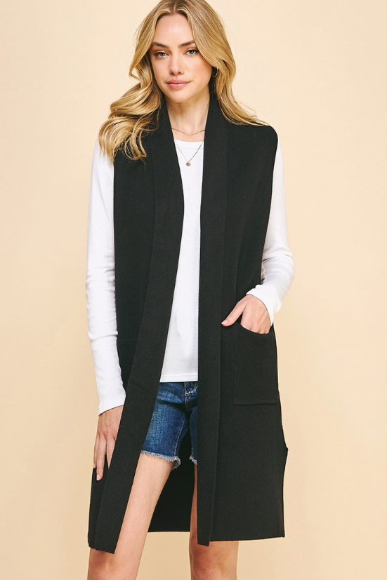 A woman modeling a long black sleeveless open-front vest over a white long-sleeve top and denim shorts, against a plain beige studio background.