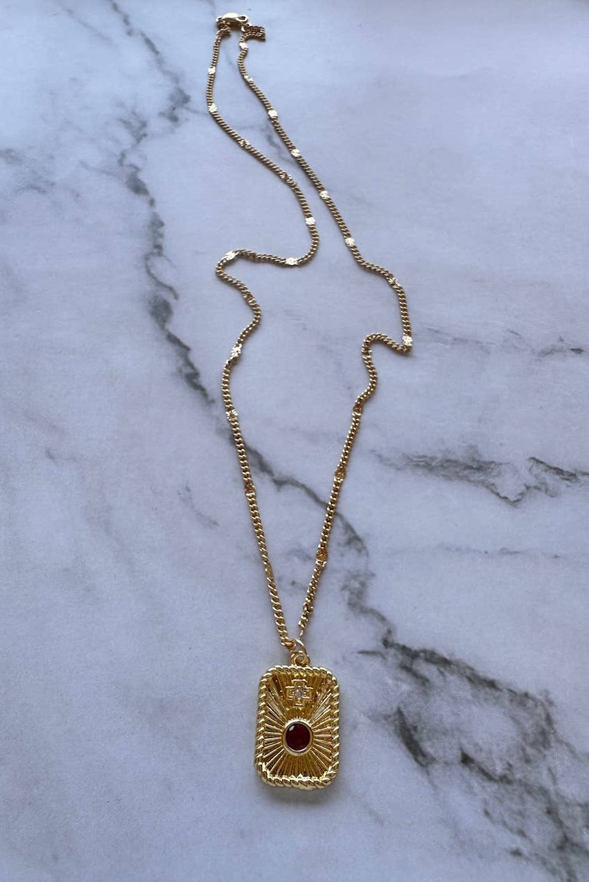 A gold chain necklace with a rectangular pendant featuring a central red gemstone and a small gold cross, displayed on a white marble surface.