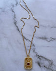 A gold chain necklace with a rectangular pendant featuring a central red gemstone and a small gold cross, displayed on a white marble surface.