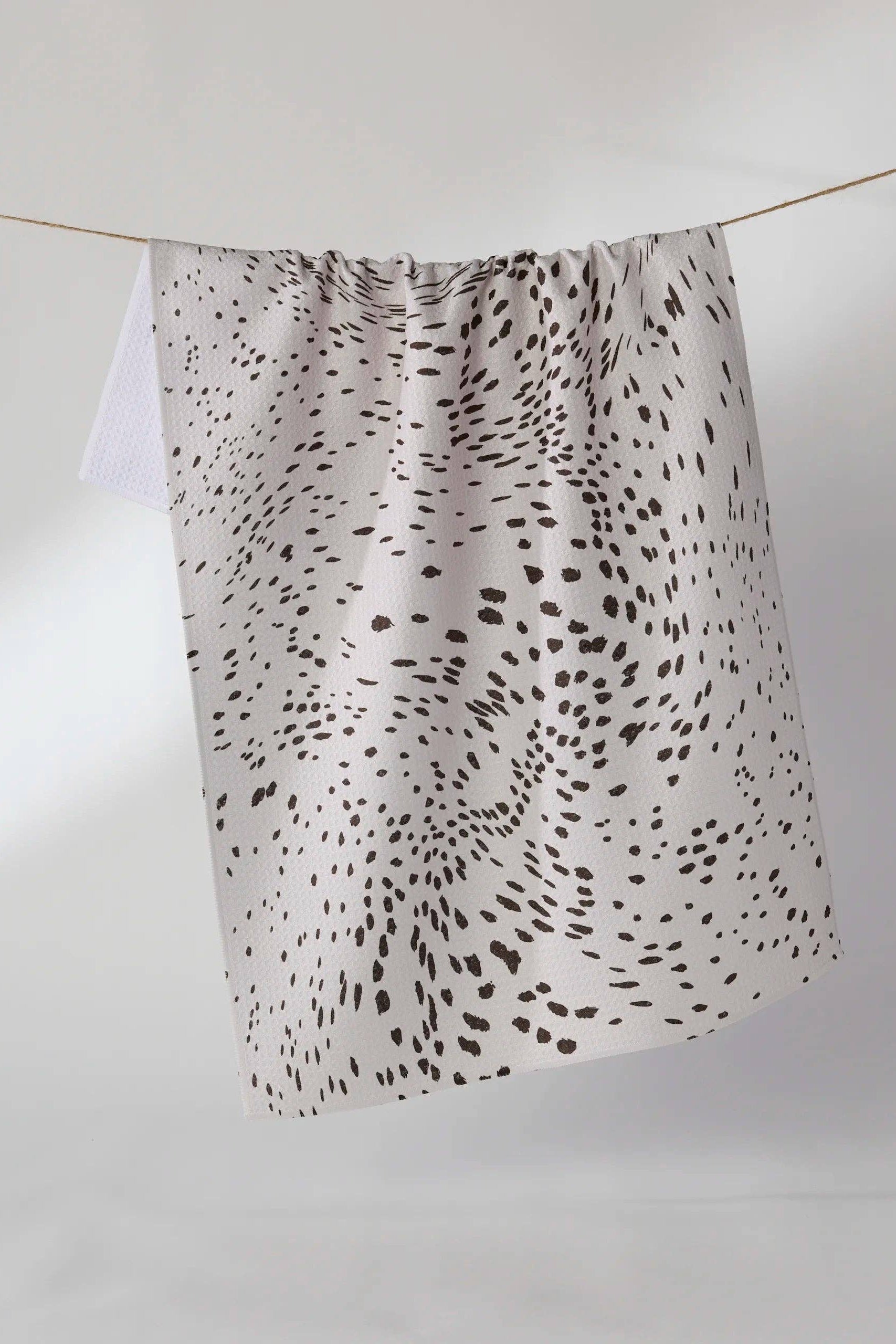 A white cotton kitchen towel with a black spotted dalmatian-style pattern, hanging from a thin wire on a plain white background.