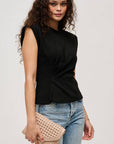 Woman wearing a black sleeveless top and blue jeans, holding a beige clutch against a plain background