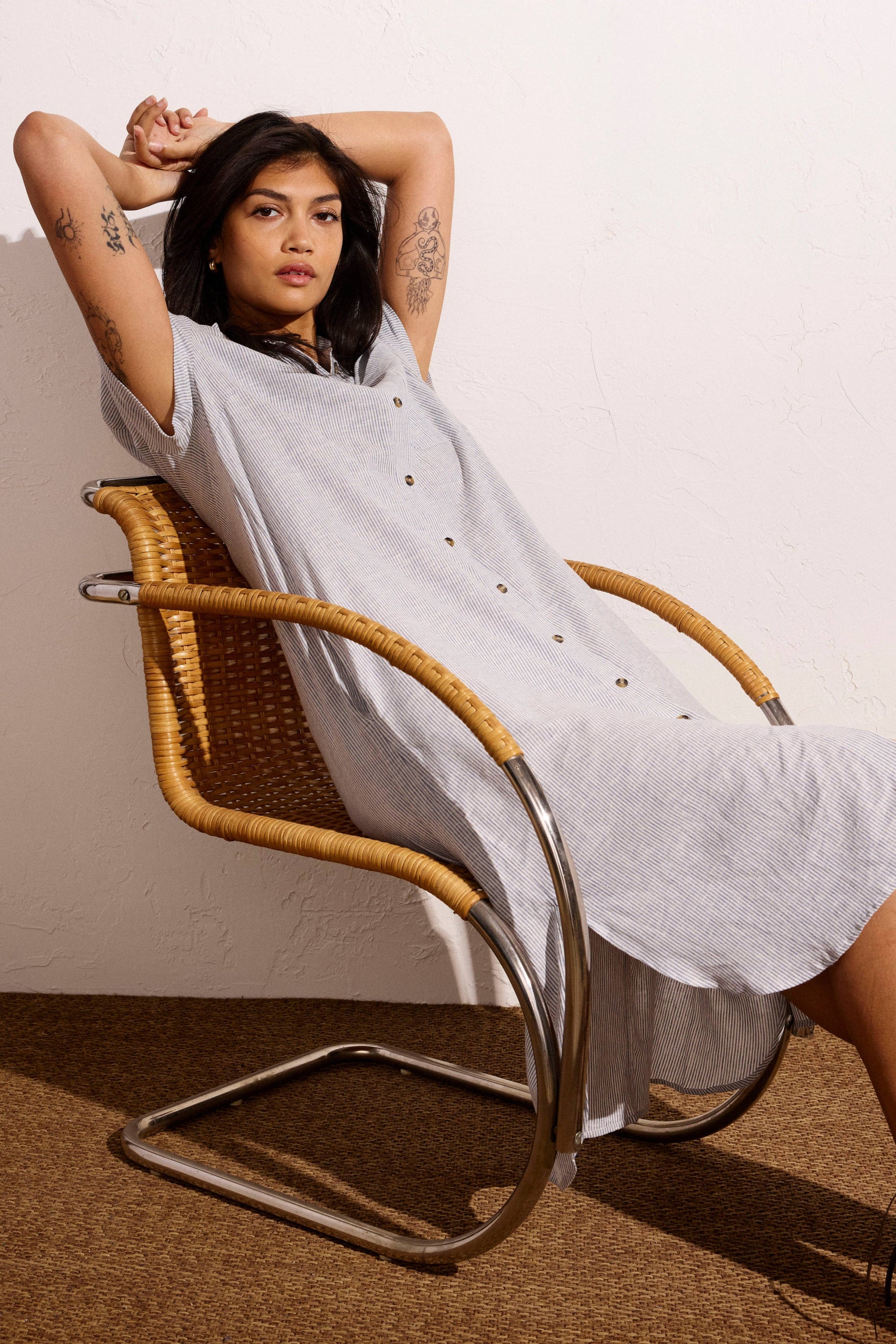 Woman in a light gray dress sitting on a wicker chair against a plain wall.