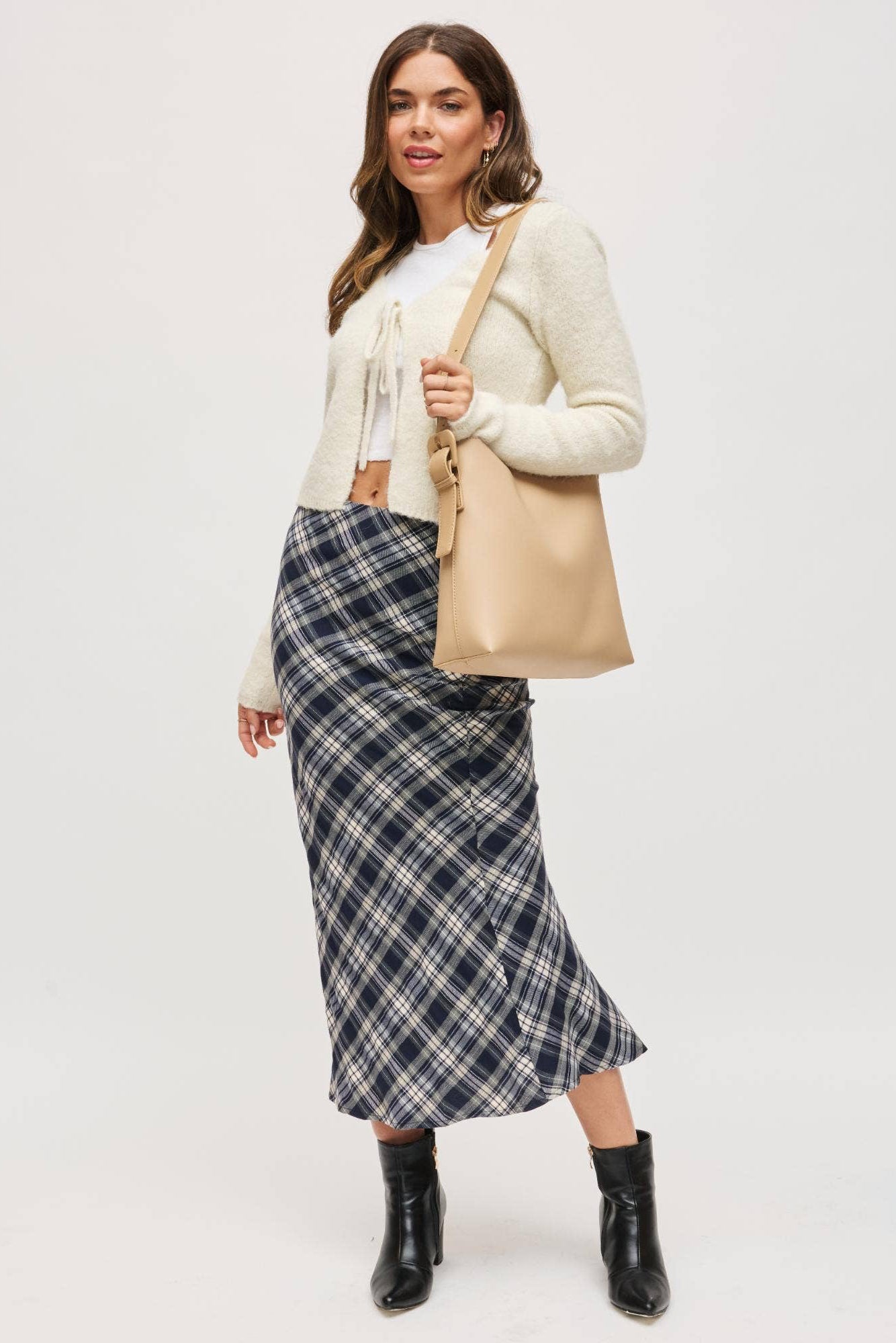 Woman wearing a plaid skirt, white top, beige cardigan, and black boots on a white background