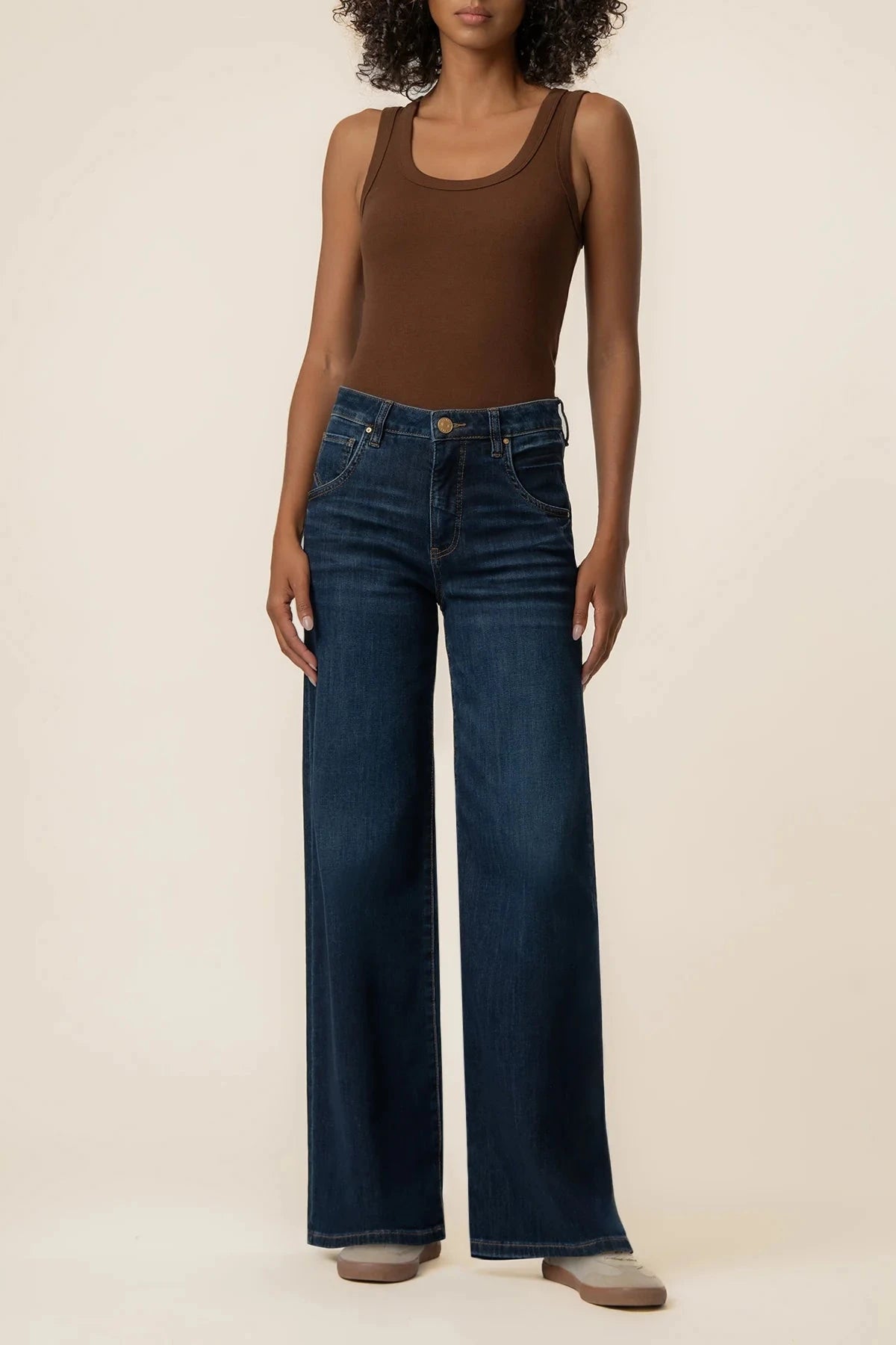 A model wearing dark blue wide-leg denim jeans and a brown ribbed tank top, standing against a solid beige studio background.
