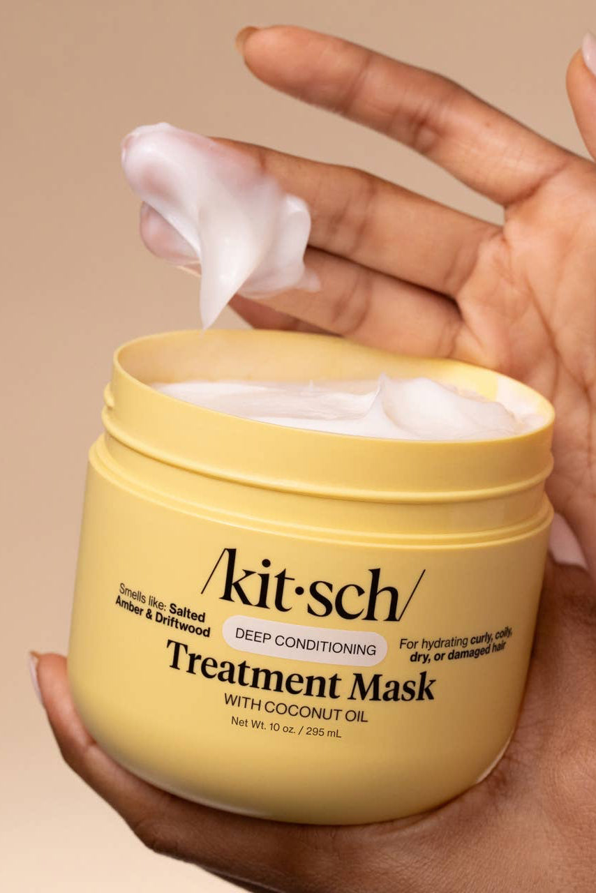 A hand holds an open yellow jar of Akit-sch Treatment Mask with coconut oil, showing white mask cream being lifted out, against a solid beige background.