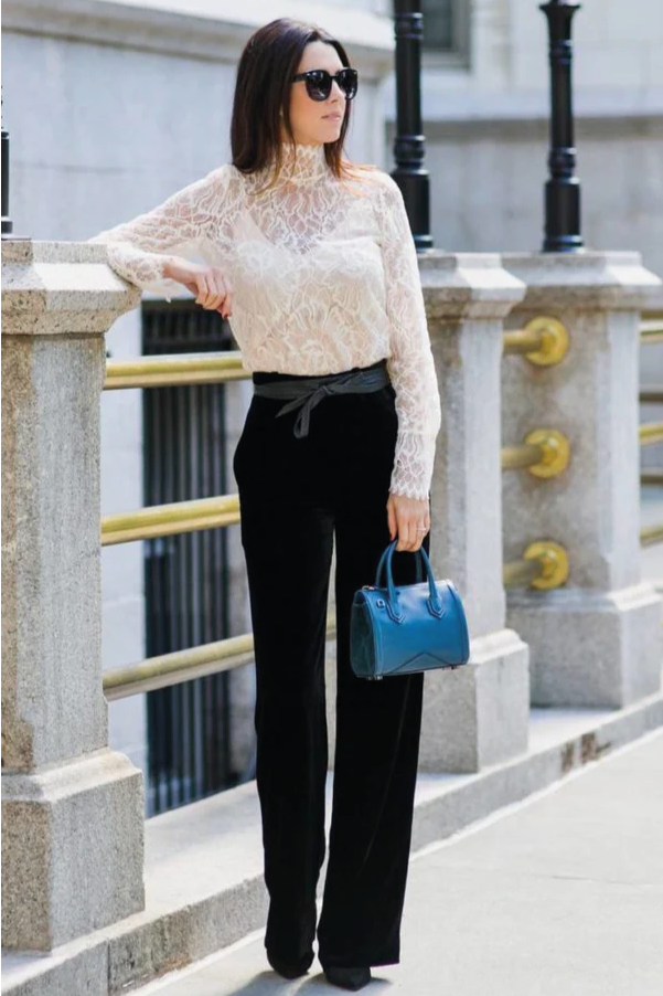 Woman in a white lace top and black pants holding a blue handbag, standing on a city street.