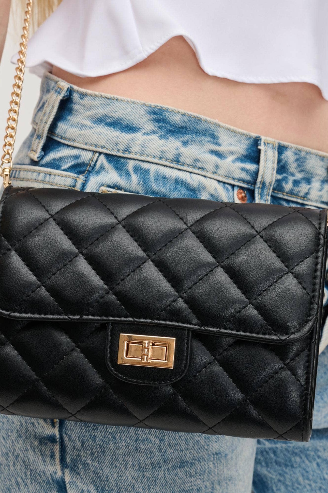 Black quilted handbag with gold chain strap held by a person wearing a white shirt and blue jeans.