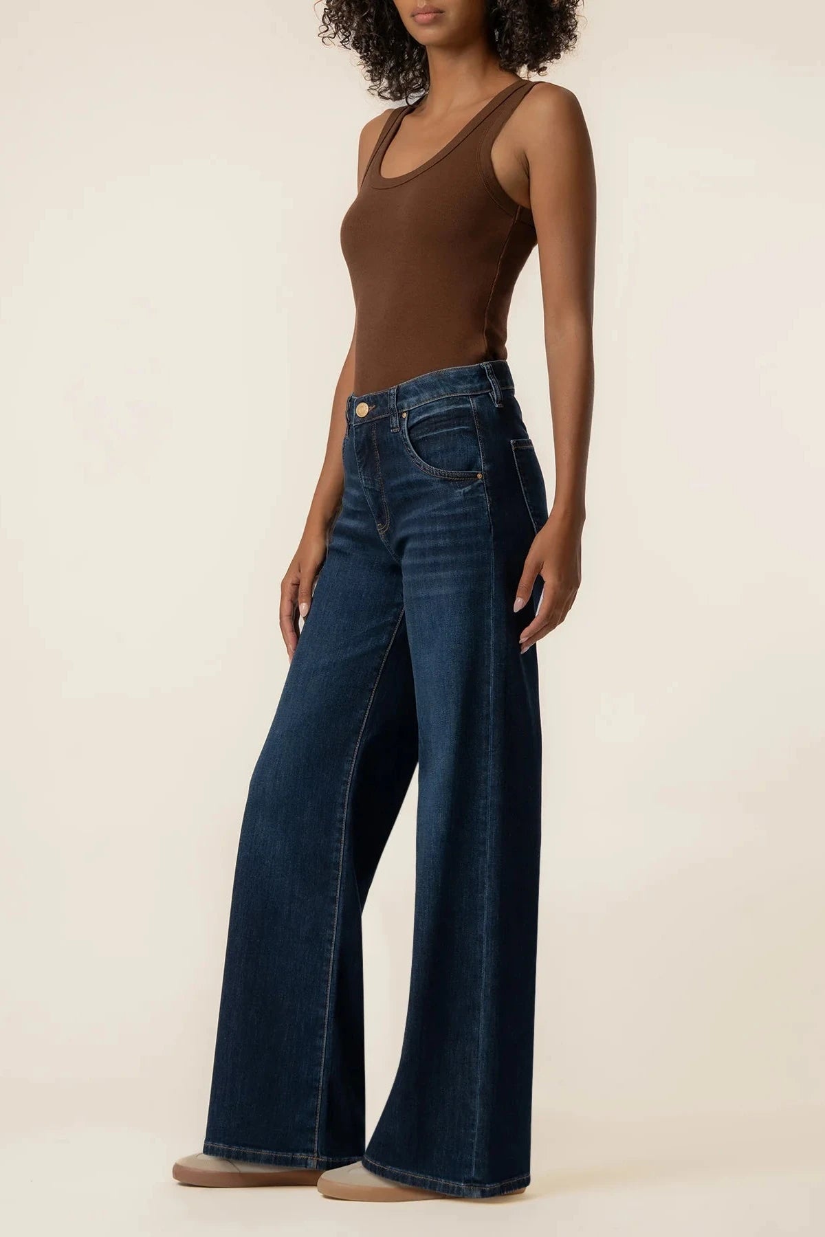 A model wearing dark blue wide-leg denim jeans and a brown ribbed tank top, standing against a plain beige studio background.