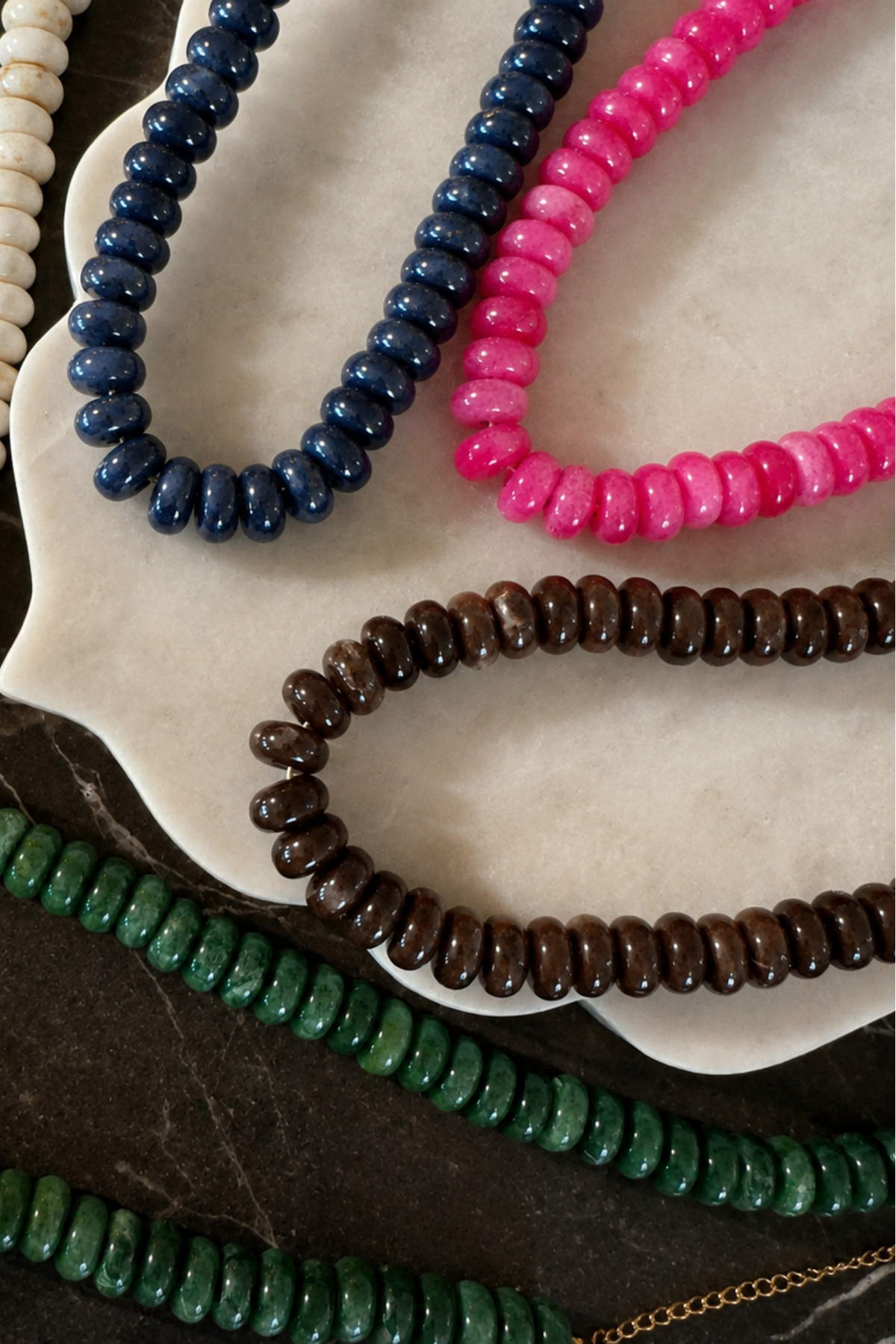 Five beaded necklaces in different colors arranged on a white scalloped dish over a dark marble surface: ivory, navy, bright pink, brown, and green. A small gold chain link is visible at the lower right.