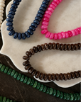 Five beaded necklaces in different colors arranged on a white scalloped dish over a dark marble surface: ivory, navy, bright pink, brown, and green. A small gold chain link is visible at the lower right.