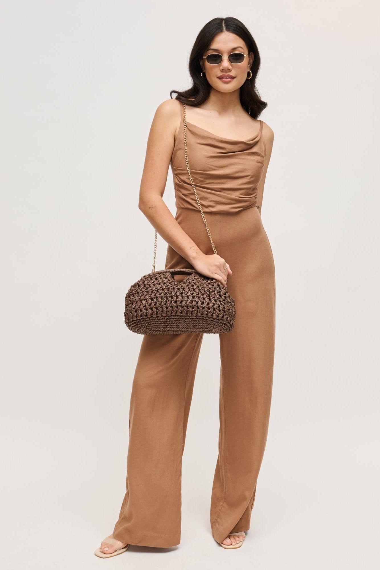 Woman in a brown jumpsuit holding a woven handbag against a plain background