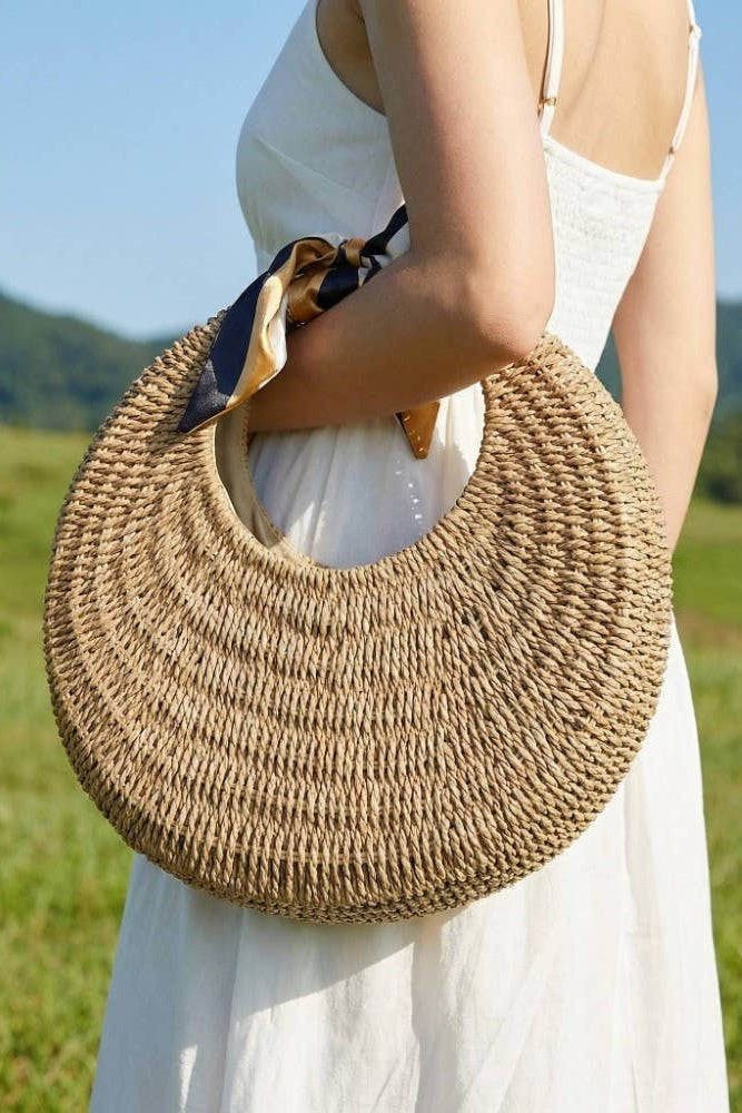 Person holding a woven bag in a scenic outdoor setting with greenery and mountains.