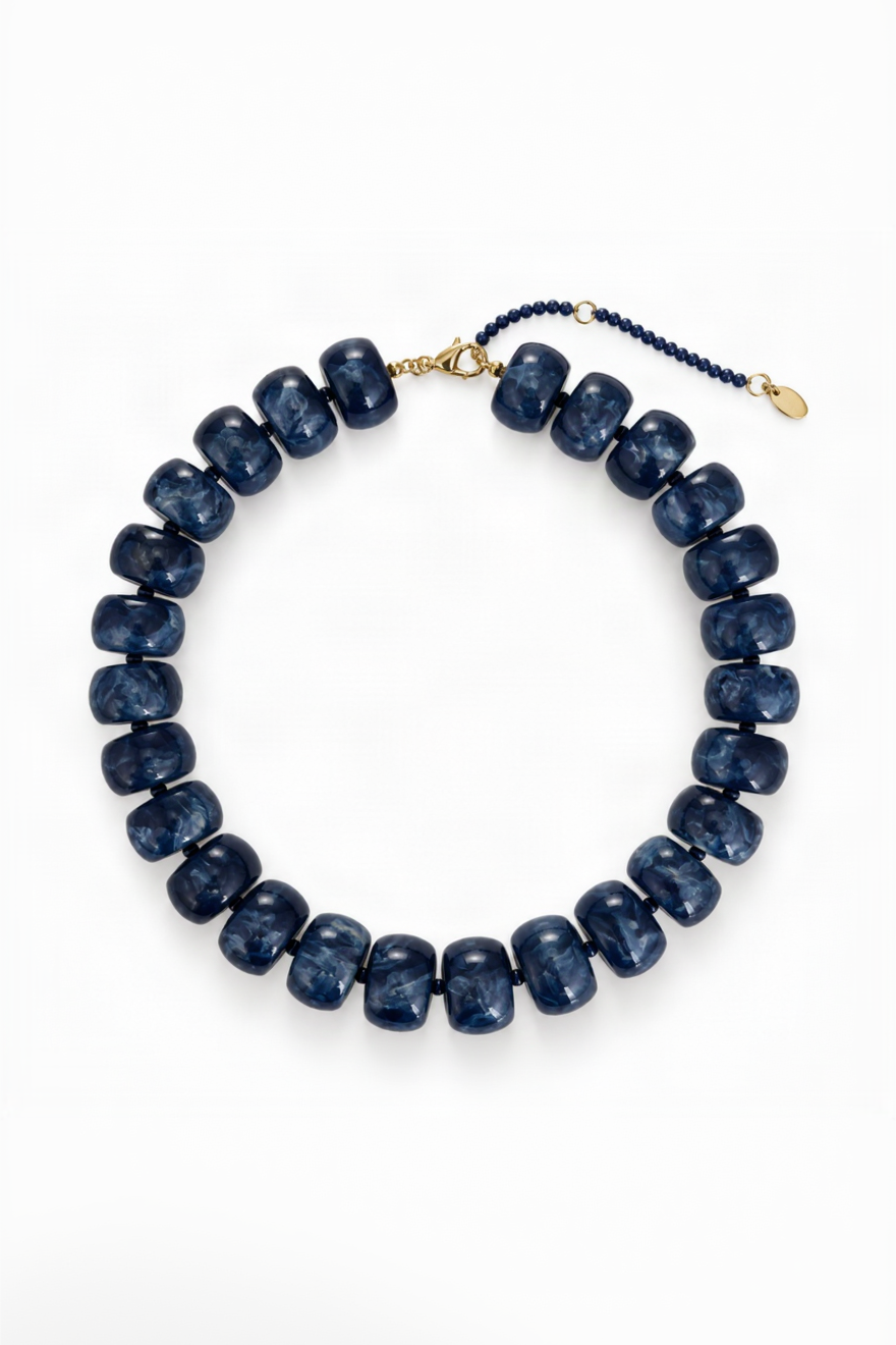 A beaded necklace made of dark blue marbled beads, arranged in a circle on a white background. It features a gold-tone clasp and a navy blue beaded extender chain with a small gold tag.