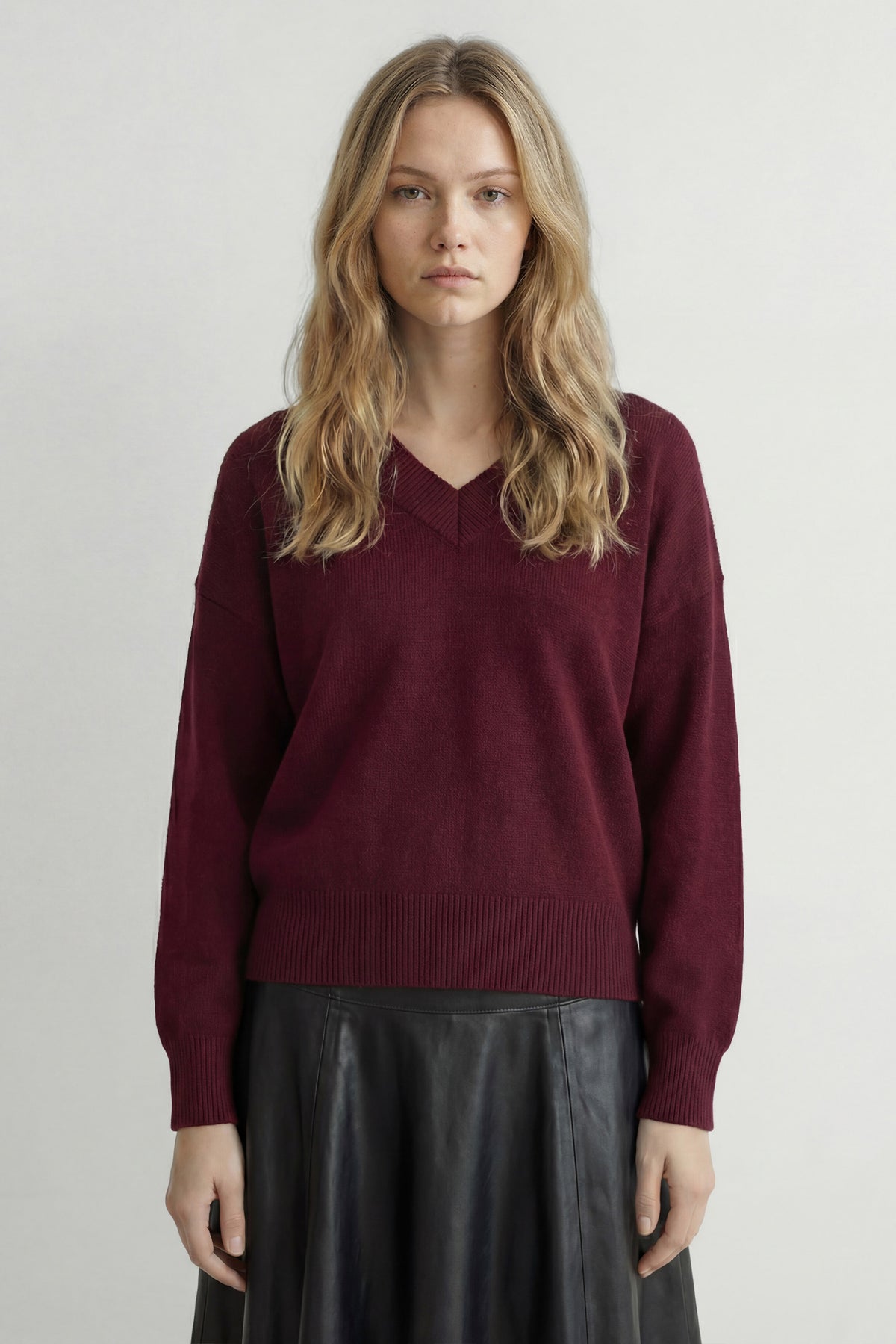 A woman wearing a burgundy knit V-neck sweater and a black skirt, standing against a light gray studio background.