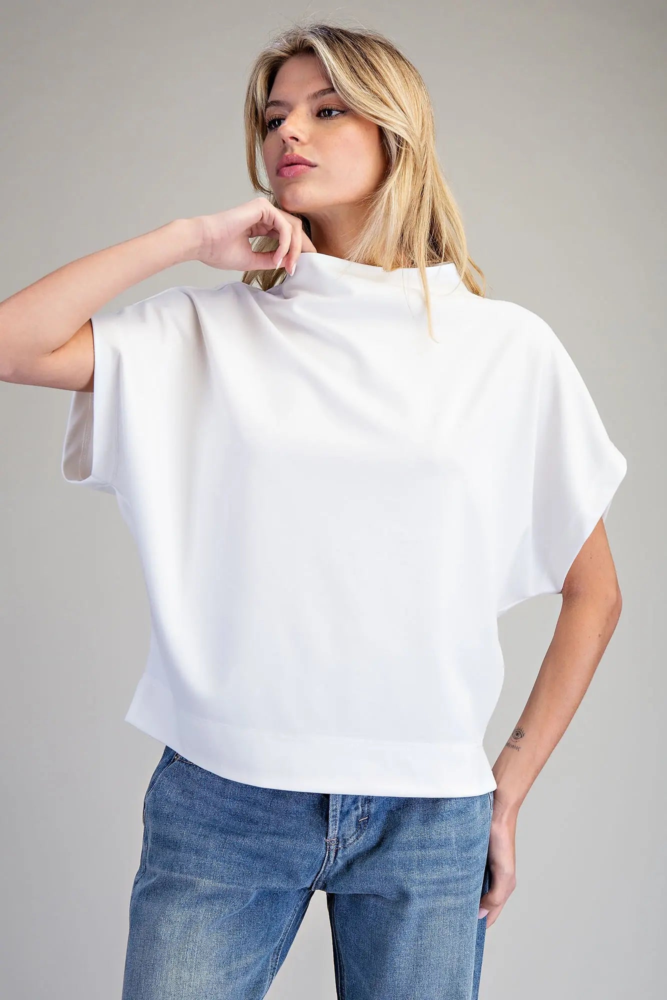 A woman wearing a white short-sleeve mock neck top and blue jeans, posing against a solid gray studio background.