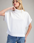 A woman wearing a white short-sleeve mock neck top and blue jeans, posing against a solid gray studio background.