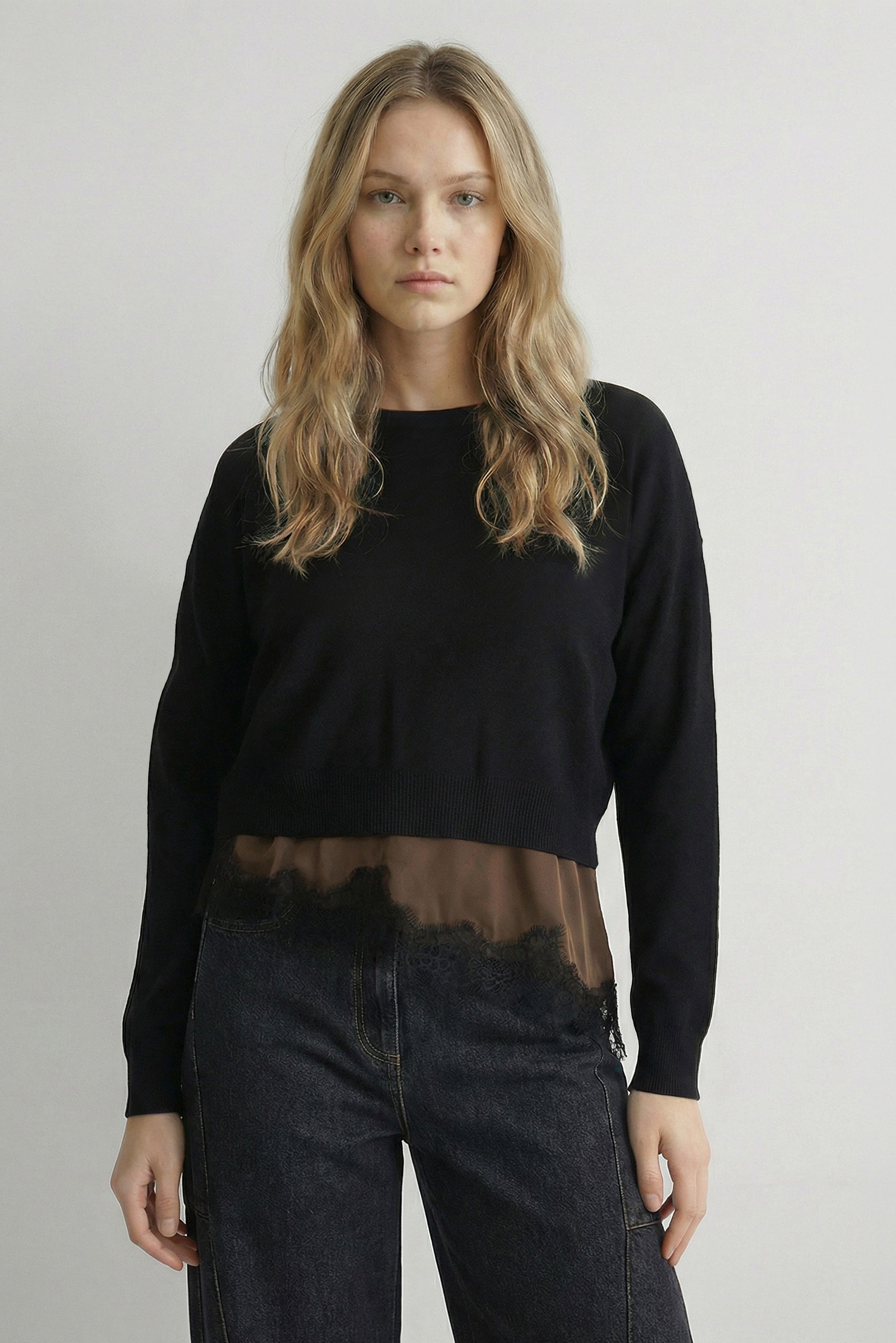 A woman modeling a black long-sleeve knit top with a sheer brown mesh band at the hem, paired with dark jeans, against a plain light gray studio background.